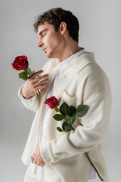 Side view of brunette man in white stylish clothes posing with red roses isolated on grey - foto de stock
