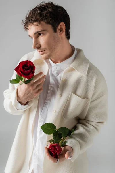 Чоловік у білій сорочці та м'якій куртці тримає червоні троянди та дивиться ізольовано на сірому — стокове фото