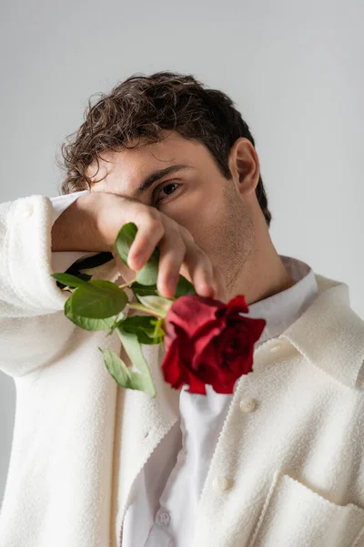 Man with red roses in sleeve of white jacket obscuring face with hand and looking at camera isolated on grey — Stock Photo