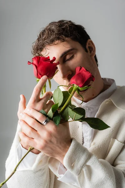 Trendy man in white clothing and silver finger ring obscuring face with red roses isolated on grey - foto de stock