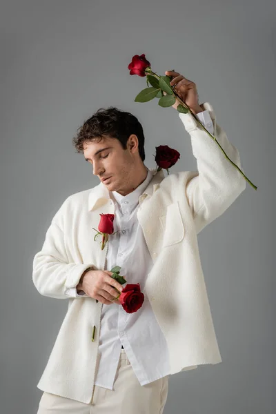 Мужчина в белой стильной одежде позирует с красными розами и закрытыми глазами, изолированными на сером — стоковое фото