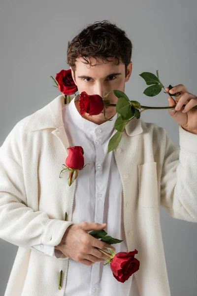 Fashionable man in white soft jacket obscuring face with red rose while looking at camera isolated on grey — Stockfoto