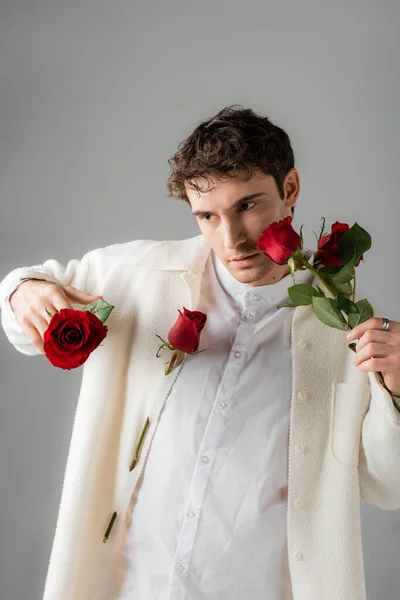 Модний чоловік в білому одязі позує з червоними і свіжими трояндами ізольовані на сірому — стокове фото
