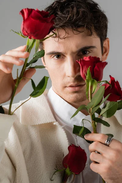 Портрет человека в белой куртке и серебряном кольце пальца, смотрящего в камеру и держащего красные розы, изолированные на сером — стоковое фото