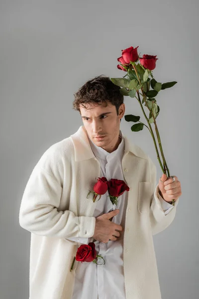 Trendy man in white soft jacket holding red roses and looking away isolated on grey — Stock Photo