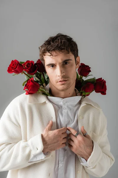 Стильний чоловік у білій сорочці і піджаку дивиться на камеру, позуючи з червоними трояндами ізольовано на сірому — стокове фото