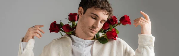 Брюнетка мужчина в белой куртке стоя с закрытыми глазами и красные розы изолированы на сером, баннер — стоковое фото