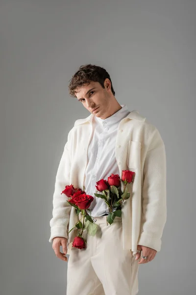 Stylish man looking at camera while posing with red roses in white trousers isolated on grey — Stock Photo
