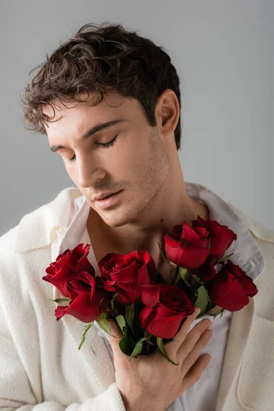Brunette man in white soft jacket standing with closed eyes and holding bouquet of red roses isolated on grey — Stock Photo
