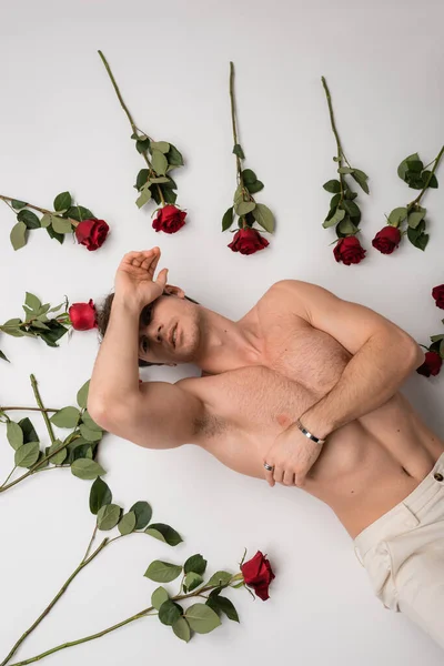 Top view of sexy shirtless man in silver bracelet lying near red roses on white background - foto de stock
