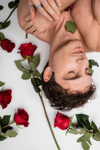 Вид зверху без сорочки в срібних кільцях пальців, що лежать із закритими очима біля червоних свіжих троянд на білому тлі — стокове фото