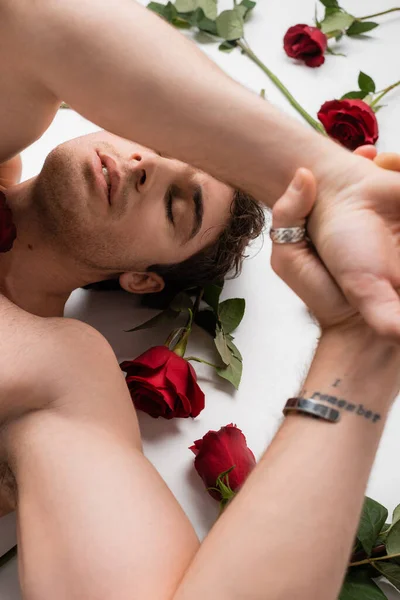 Top view of tattooed shirtless man lying with hands above head near red roses on white background - foto de stock