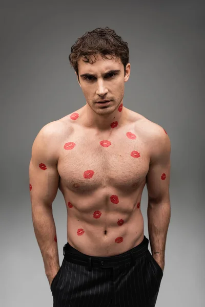 Shirtless man with red kisses on muscular body standing with hands in pockets of black pants on grey background - foto de stock