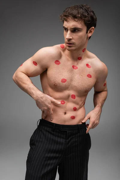 Sexy man in black pants touching muscular torso with red lipstick marks and looking away on grey background — стоковое фото