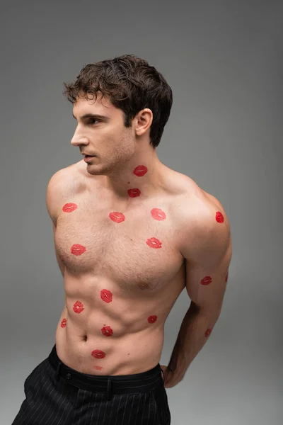 Брюнетка мужчина с красными отпечатками поцелуя на туловище без рубашки глядя в сторону на сером фоне — стоковое фото