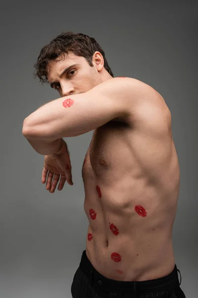 Sexy shirtless man with red kiss prints on body obscuring face with arm while looking at camera isolated on grey — Foto stock