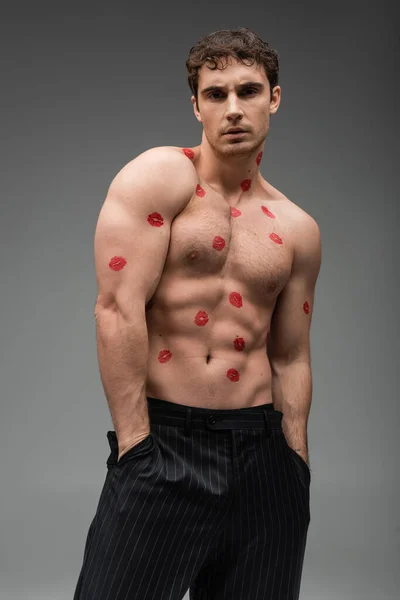 Sexy athletic man with red kiss prints on body standing with hands in pockets of black trousers on grey background — Stock Photo