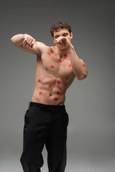 Athletic man with red kisses on shirtless body obscuring face while posing with closed eyes on grey background — Stock Photo