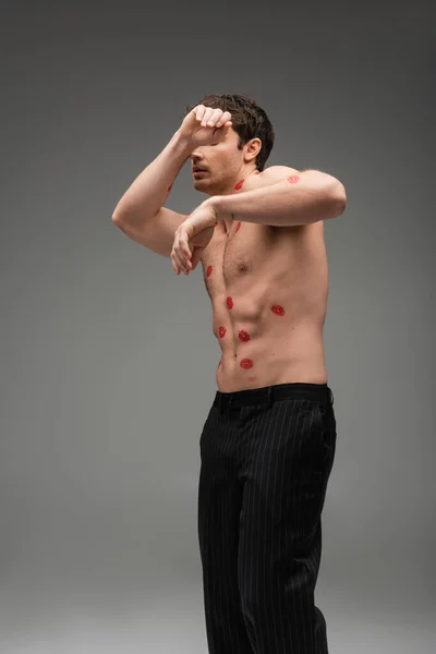 Man in black pants with red lip prints on shirtless torso obscuring face while posing on grey background - foto de stock
