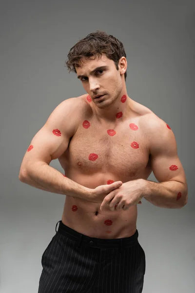 Атлетичный мужчина с красными отпечатками губ на мускулистом теле, смотрящий на камеру на сером фоне — стоковое фото