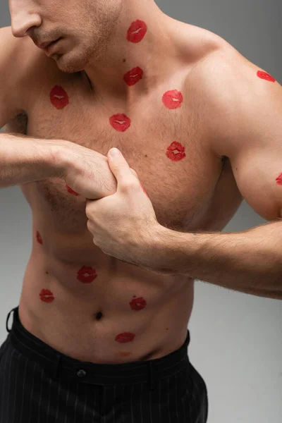 Частичный вид человека без рубашки с красными отпечатками поцелуев на мускулистом теле, изолированном на сером — стоковое фото