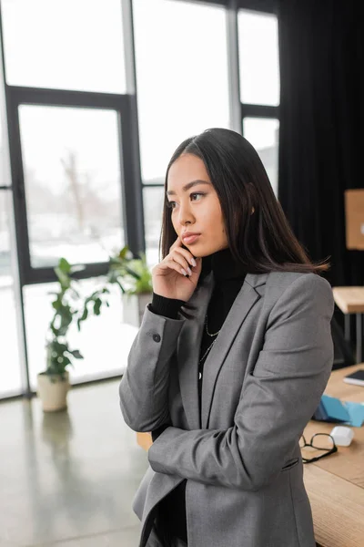 Pensive asian designer in blazer looking away while standing in office — Stock Photo