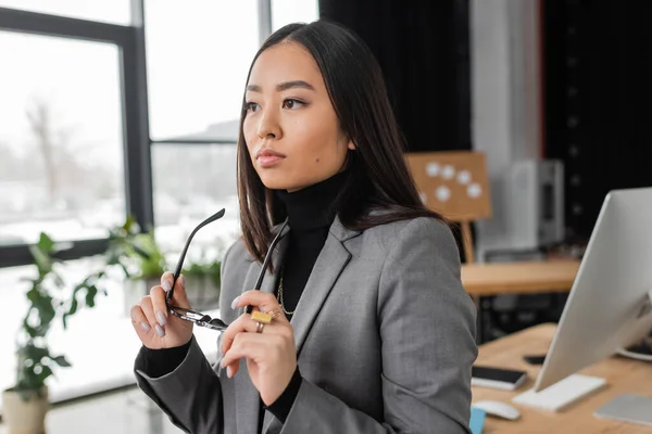 Pensive and young asian designer holding eyeglasses in office - foto de stock