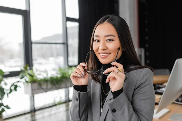 Позитивный азиатский дизайнер держит очки и улыбается в офисе перед камерой — стоковое фото