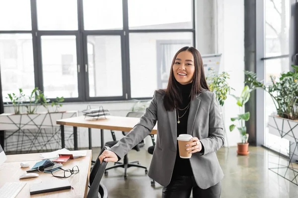 Positive asian interior designer holding coffee to go and looking at camera in office — Stock Photo