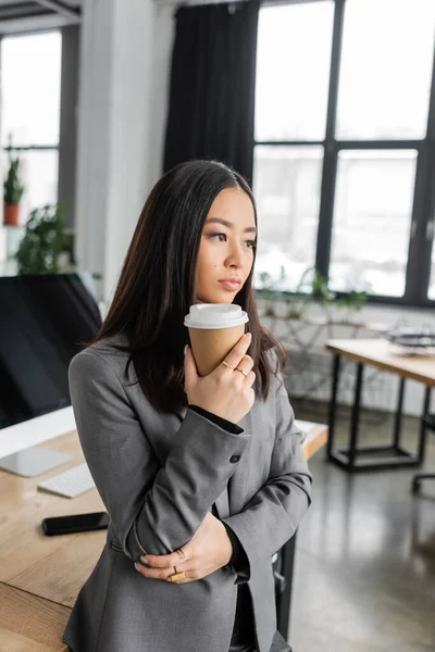 Brunette asian interior designer holding paper cup while thinking in office — Stock Photo