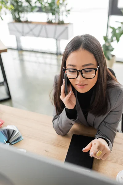 High angle view of asian interior designer talking on smartphone and pointing at computer in office — Foto stock