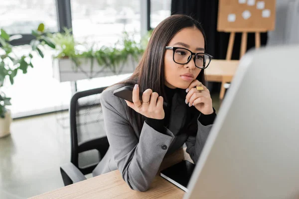 Asian designer holding smartphone and looking at computer monitor in office — Stockfoto