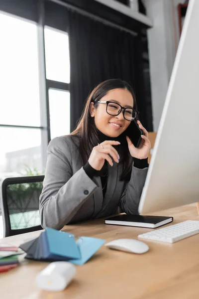 Smiling asian interior designer talking on smartphone near computer and color samples — Photo de stock