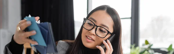 Asian designer talking on smartphone and holding blurred color samples in studio, banner — Fotografia de Stock