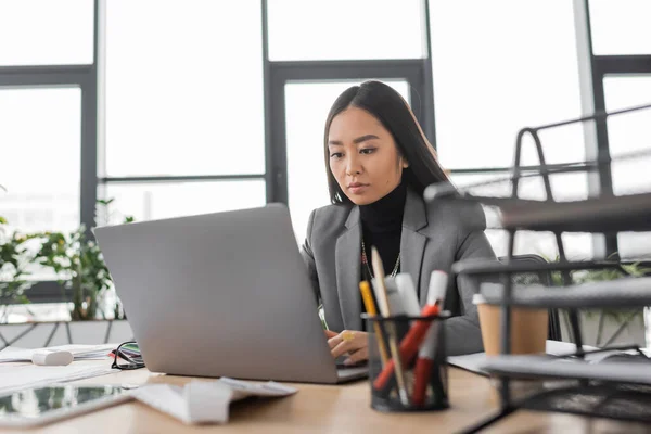Brunette asian designer using laptop near digital tablet on desk in office — Stockfoto