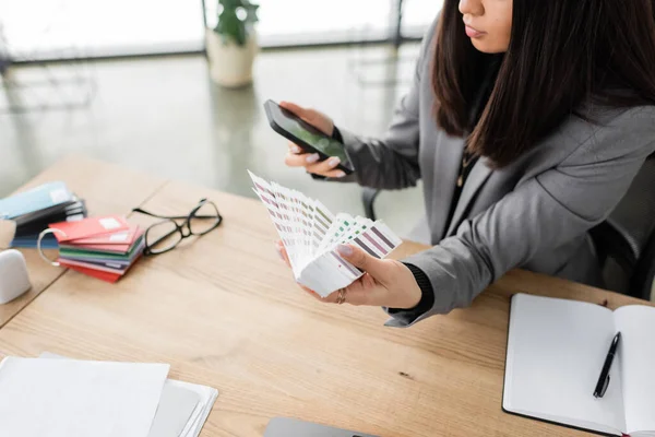 Cropped view of interior designer using smartphone and holding color samples in office — Stock Photo