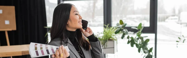 Brunette asian designer talking on cellphone and holding color samples in office, banner - foto de stock