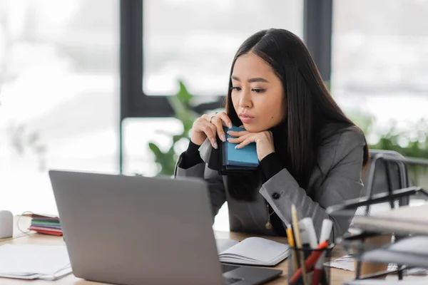 Брюнетка азиатский дизайнер держа образцы цвета и глядя на размытый ноутбук в офисе — стоковое фото
