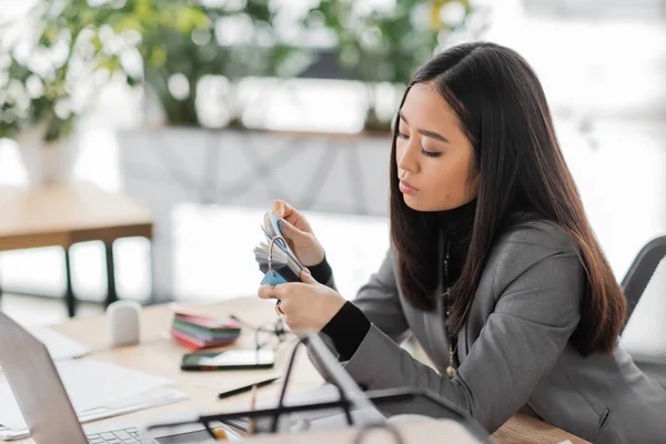 Brunette asian interior designer looking at color samples near gadgets in office — Stock Photo