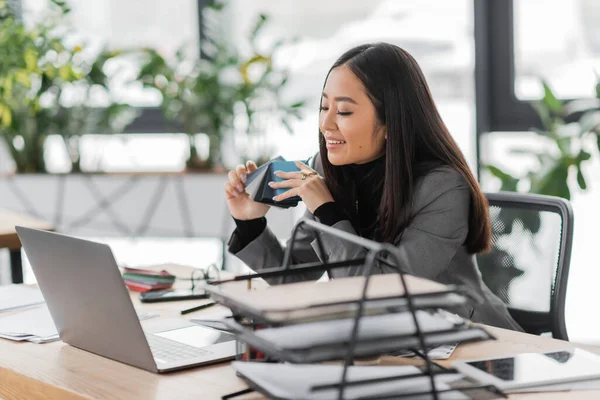 Cheerful asian interior designer looking at laptop and holding color samples in office - foto de stock