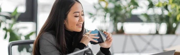 Smiling asian designer holding color swatches looking at laptop in studio, banner — Stock Photo