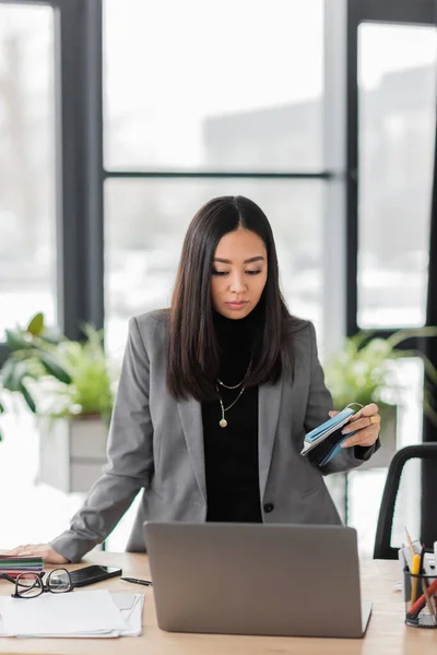Азиатский дизайнер интерьеров держит цветные образцы возле ноутбука и очков на рабочем столе — стоковое фото