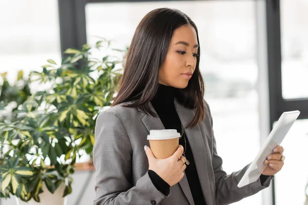 Brunette asian interior designer holding paper cup and using digital tablet in studio — Foto stock