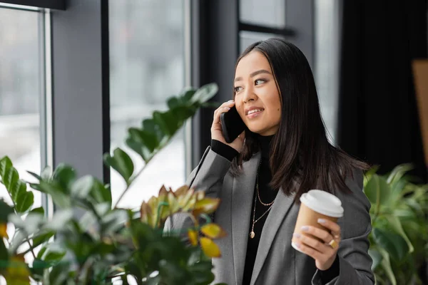 Smiling asian designer talking on smartphone and holding paper cup near plants in studio — Stock Photo