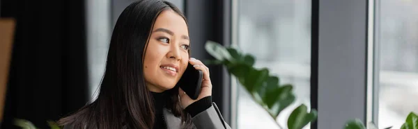 Positive asian designer talking on mobile phone in office, banner — Stock Photo