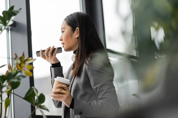 Side view of asian interior designer recording voice message on smartphone and holding coffee in studio — Stock Photo