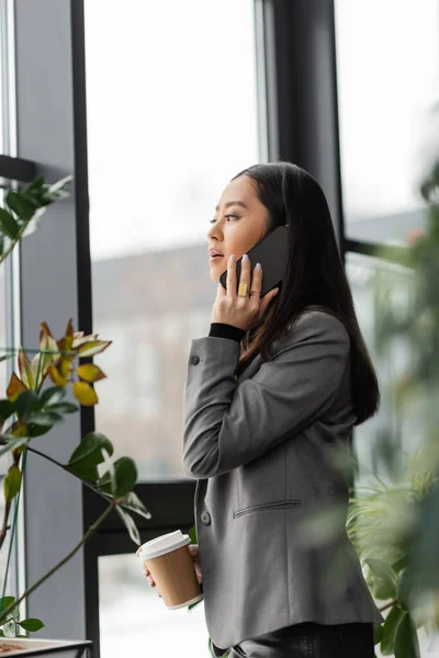 Asian designer talking on smartphone and holding takeaway coffee in office — Stock Photo