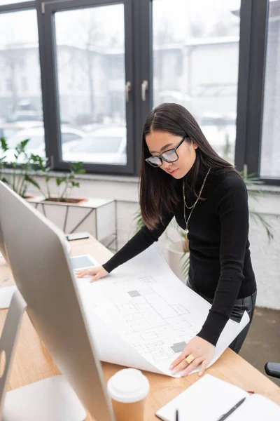 Brunette asian designer looking at blueprint near computer and coffee in studio — Stock Photo