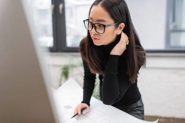 Brunette asian interior designer looking at computer near blurred blueprint in office — Stockfoto