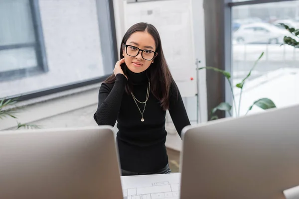 Young asian designer in eyeglasses looking at camera near computers and blueprint in office — Foto stock